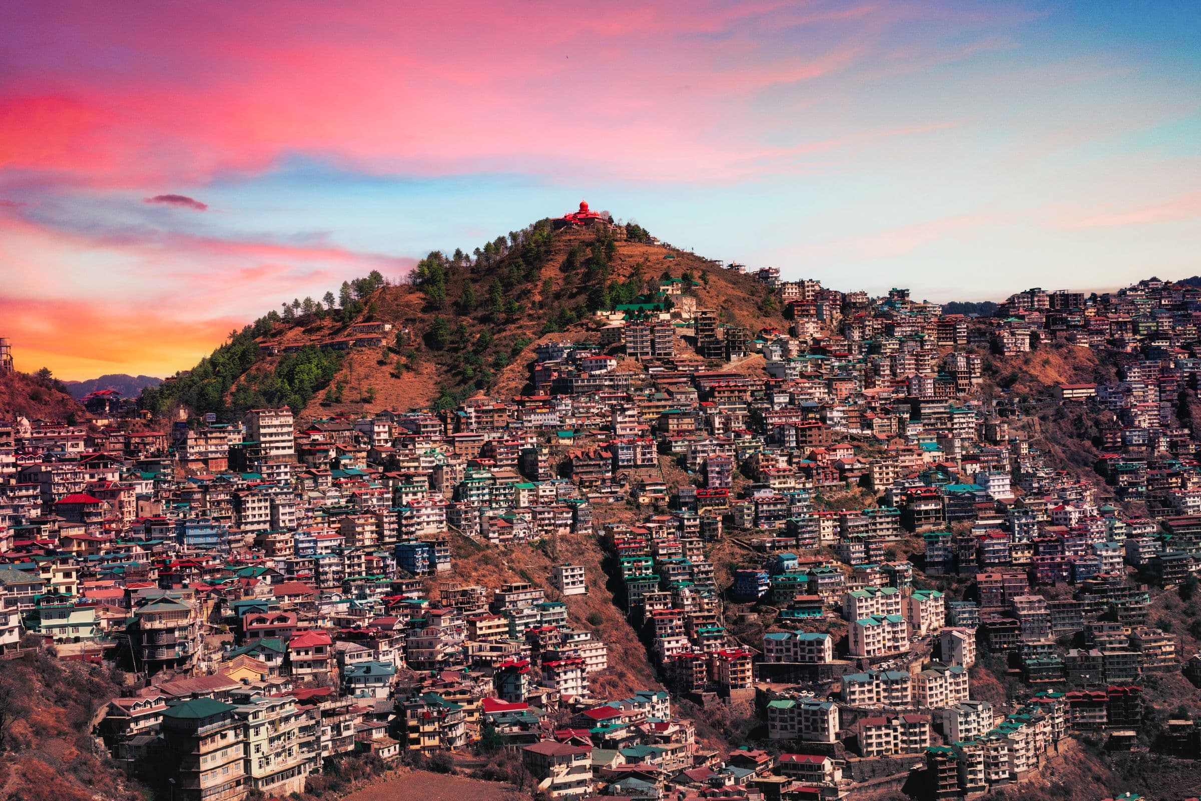 Shimla at sunset: houses with red and green roofs packed down a hillside under a pink sky.